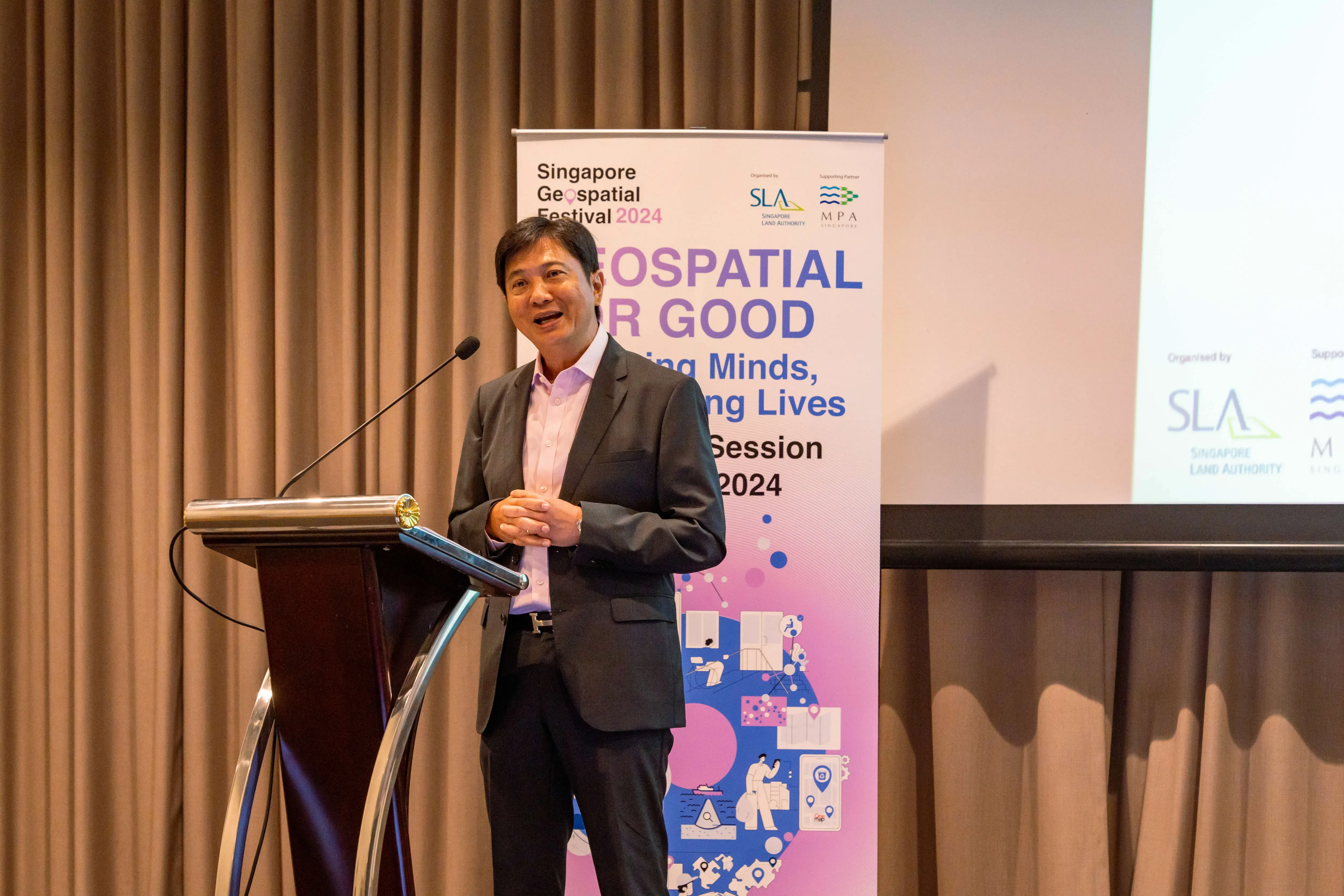 Man in a dark suit speaking at a podium beside a Singapore Geospatial Festival 2024 banner.